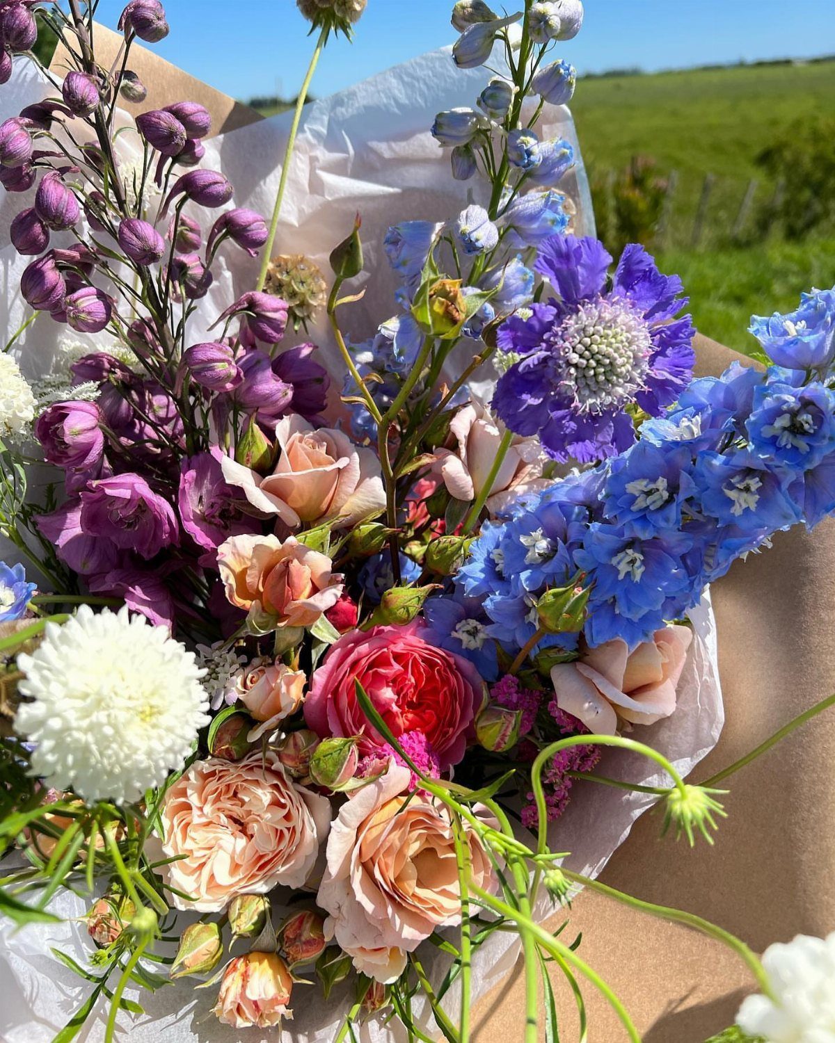Colorful bouquet of roses and delphiniums wrapped in brown paper outdoors.
