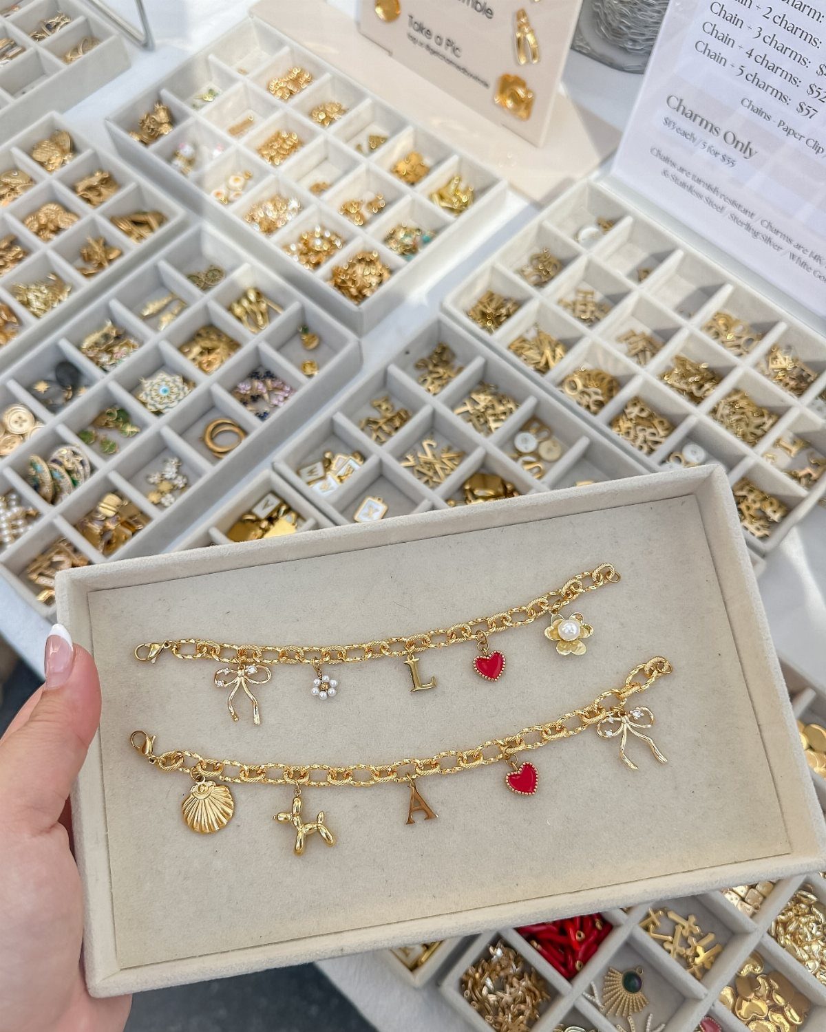 Two gold charm bracelets in a beige tray surrounded by jewelry displays