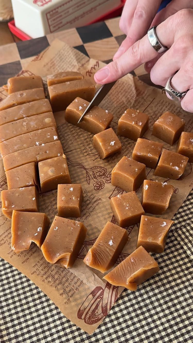 Hands cutting salted caramel fudge squares on parchment paper over a checkered cloth.