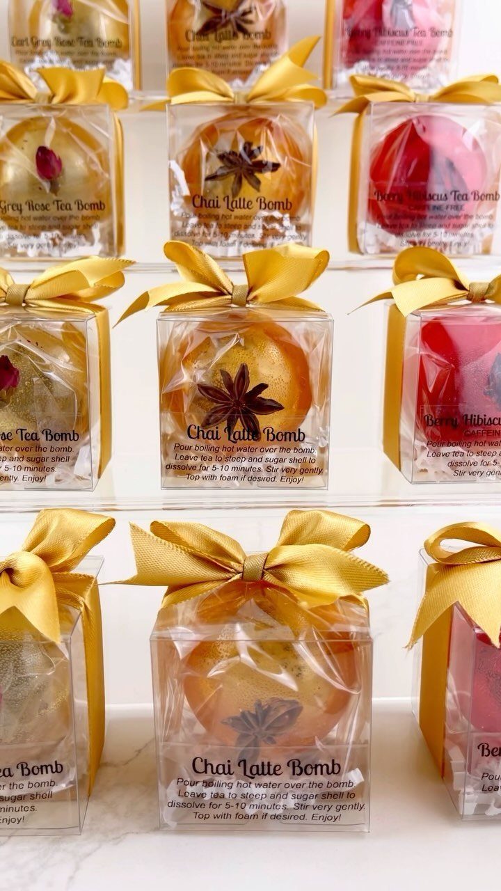 Colorful tea bombs in clear gift boxes tied with gold ribbons on white background