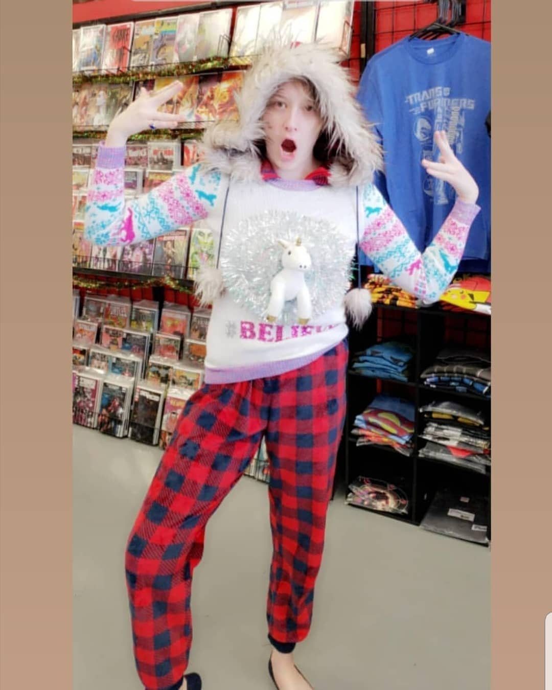 Person wearing a white unicorn holiday sweater and red plaid pajama pants posing indoors.