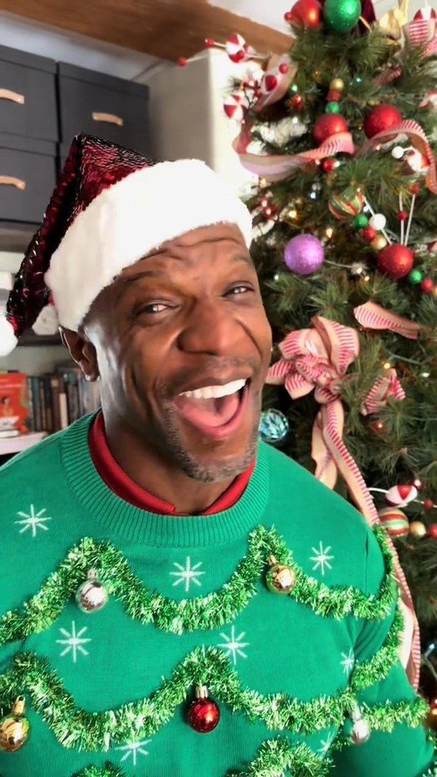 Man in a festive green ugly Christmas sweater and Santa hat smiling by a tree