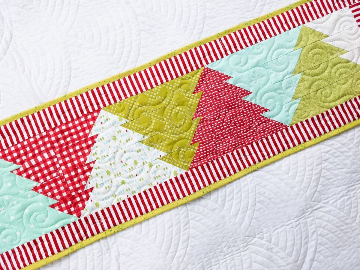 Quilted table runner with colorful Christmas tree motifs and red striped borders.