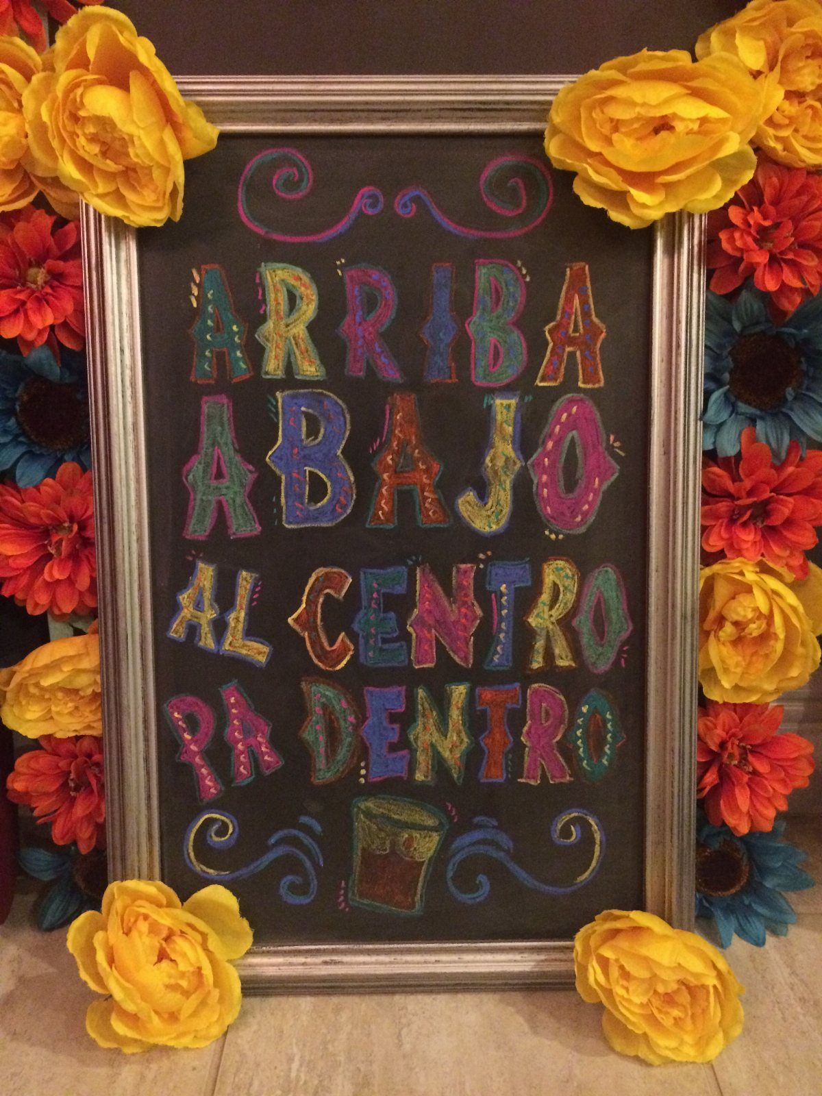 Framed chalkboard with colorful Spanish text surrounded by bright yellow and red flowers.