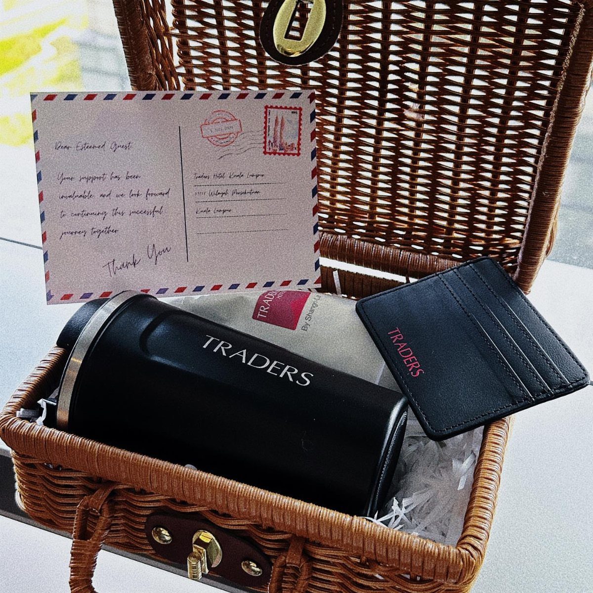 Wicker gift basket with branded black thermos, leather card holder, and handwritten letter.