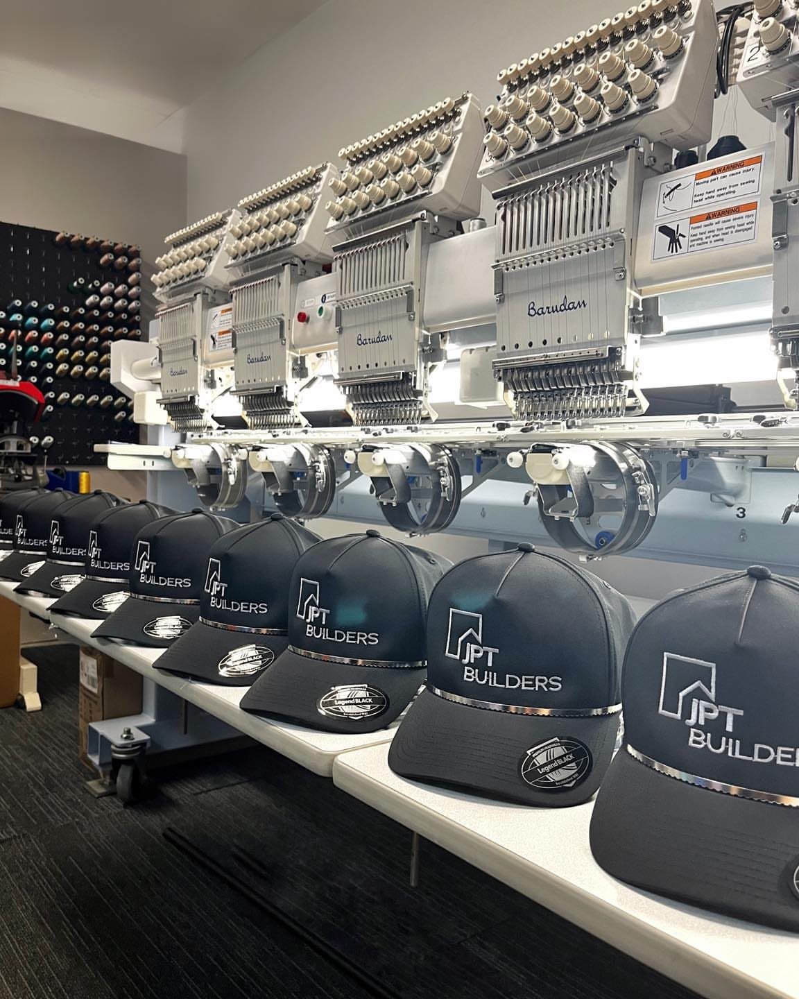 Dark grey baseball caps with company logos lined up under embroidery machines.