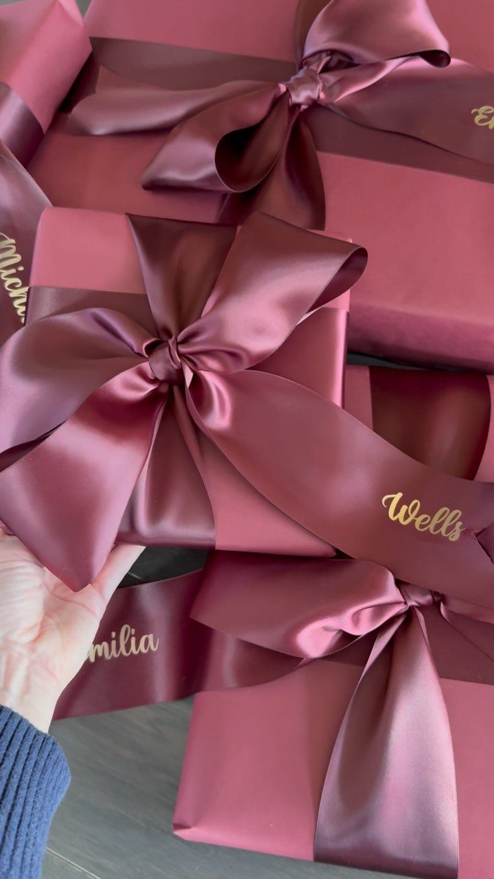 Mauve wrapped gift boxes with satin bows and personalized gold names on ribbons.