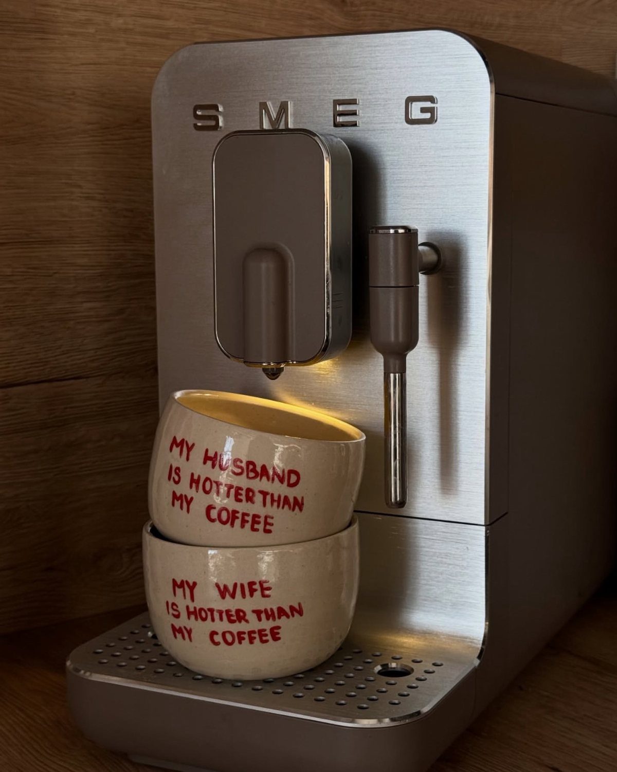 Two white ceramic mugs with red text stacked under a silver espresso machine.