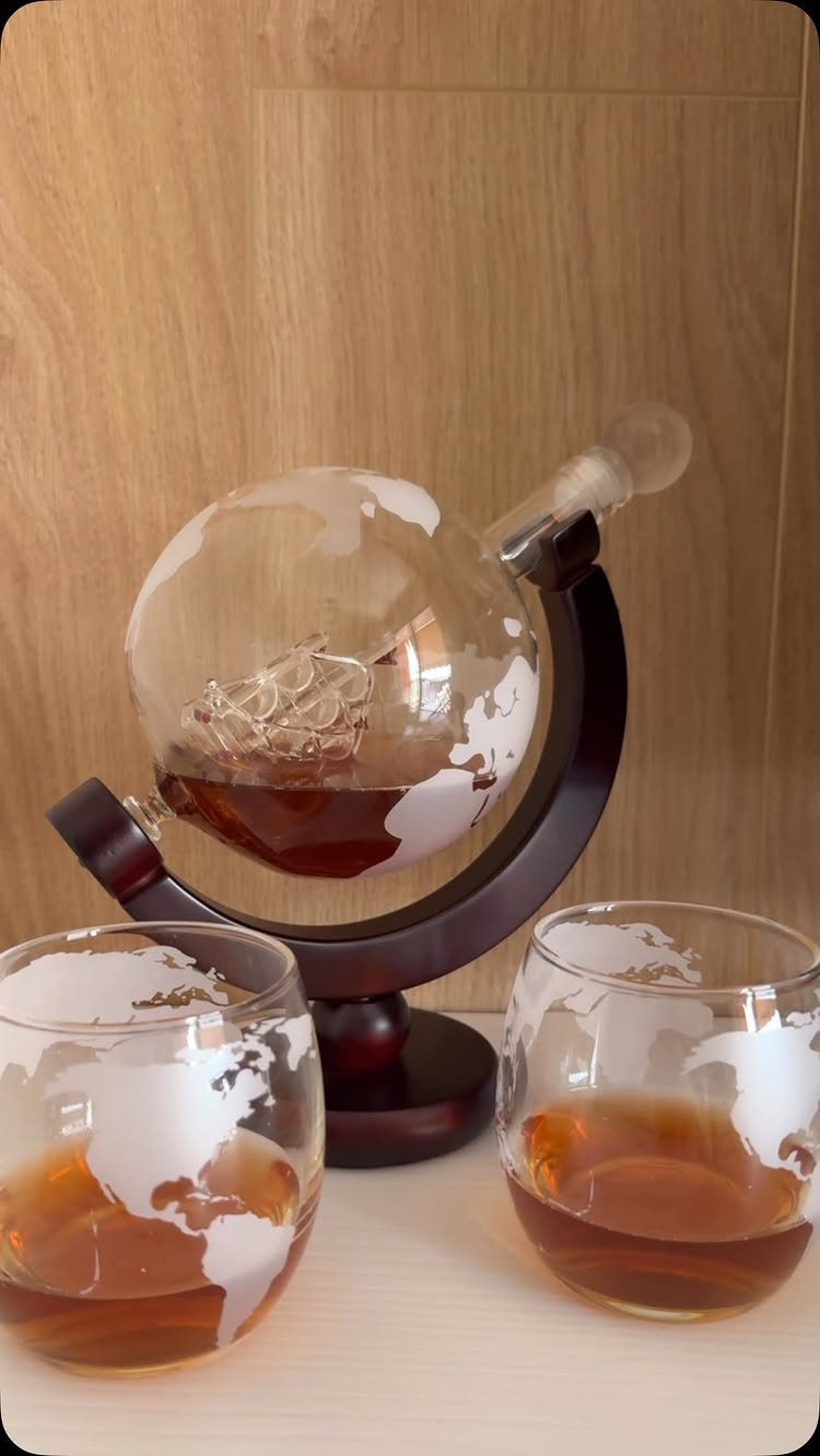 Globe-shaped glass decanter and two matching glasses filled with amber liquor on wood.