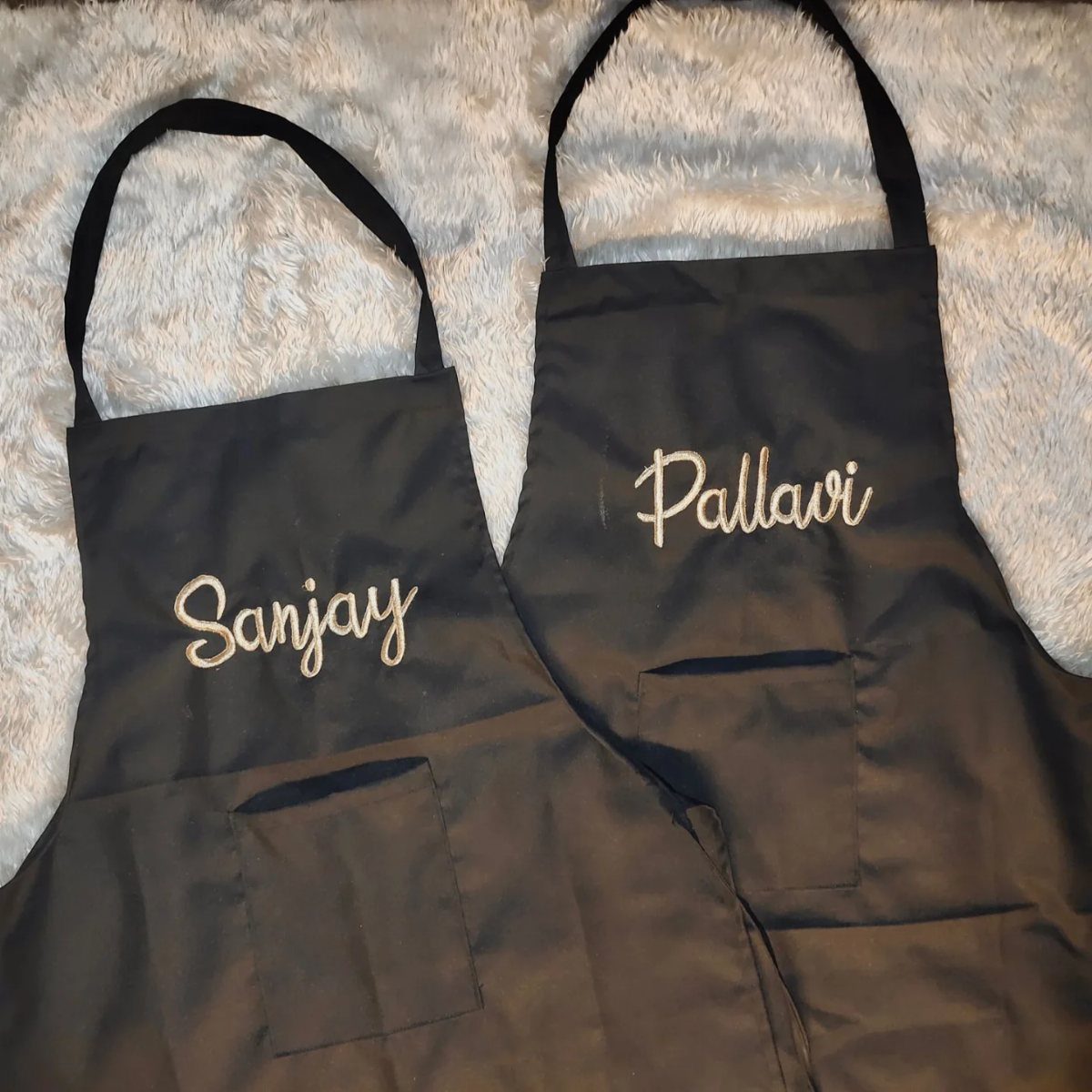 Two black aprons with the names Sanjay and Pallavi embroidered in gold cursive.