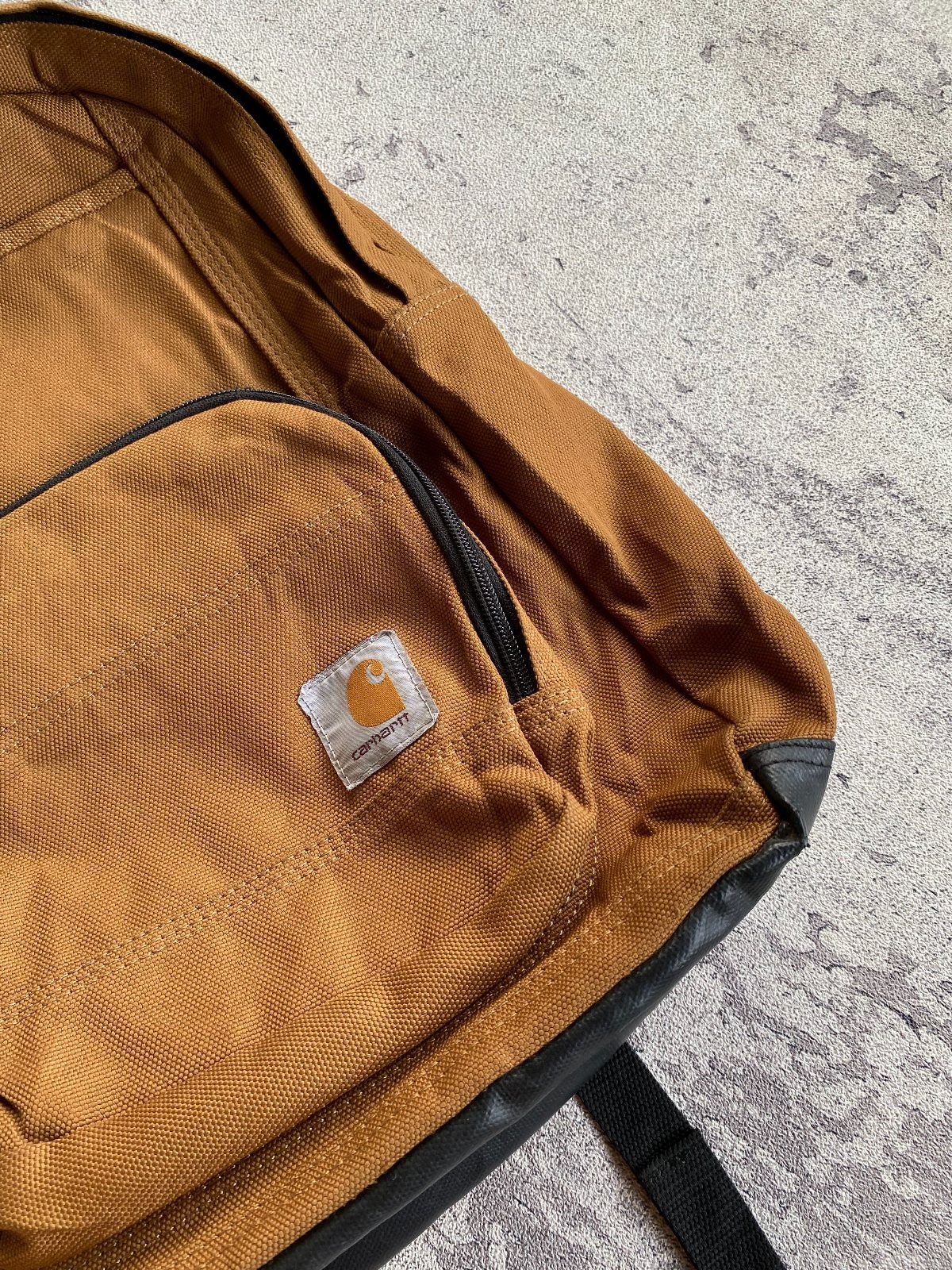 Brown canvas Carhartt backpack resting on a grey concrete ground.