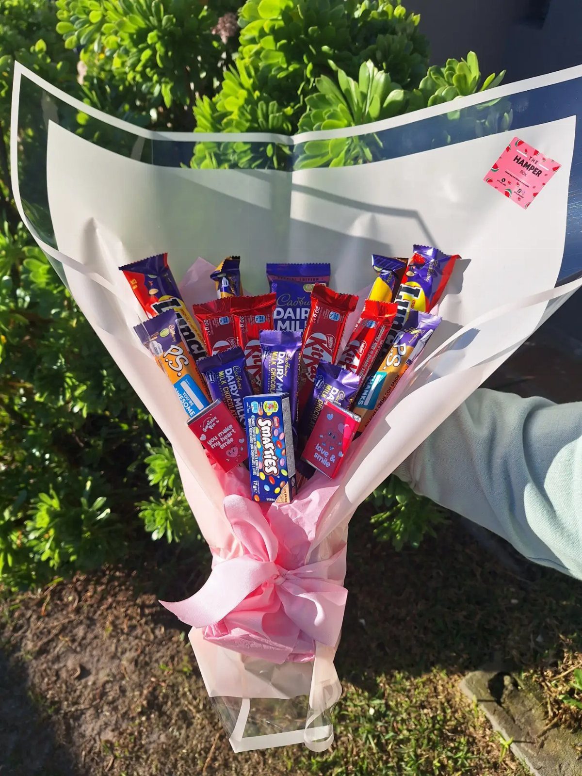 Candy bouquet wrapped in white paper and pink ribbon against green bushes.