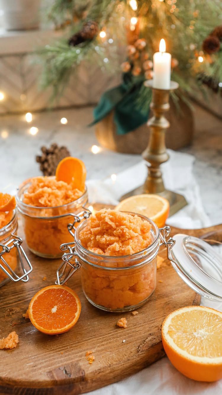 Orange body scrubs in glass jars on a wooden board with citrus slices.