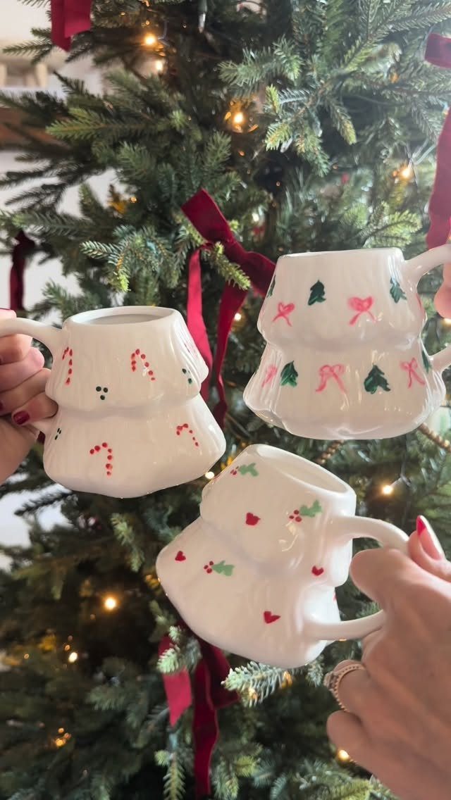 Hand-painted white Christmas mugs held in front of a decorated holiday tree.