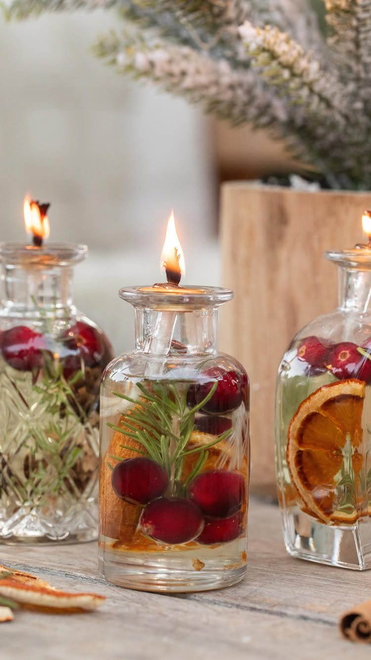 Three glass bottle candles filled with cranberries, rosemary, and orange slices, lit on wood.