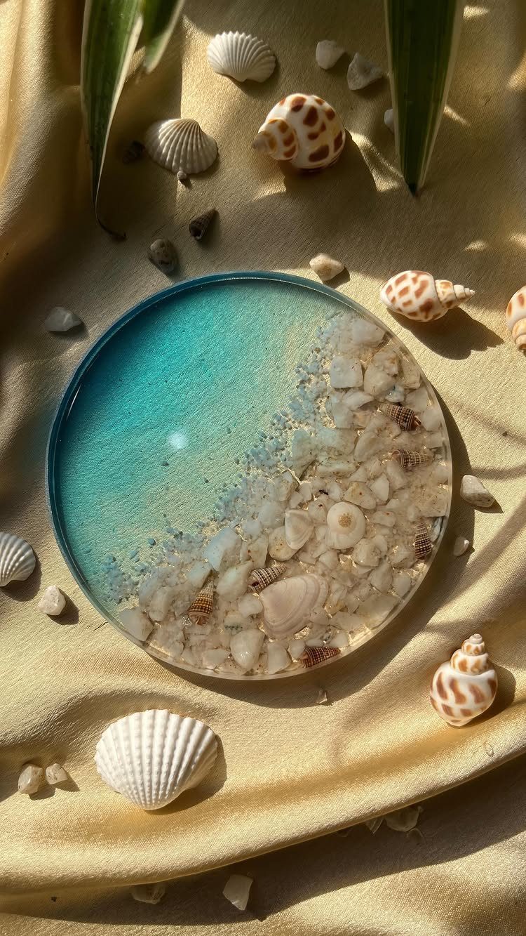 Circular resin coaster with blue ocean colors and embedded shells on beige fabric.