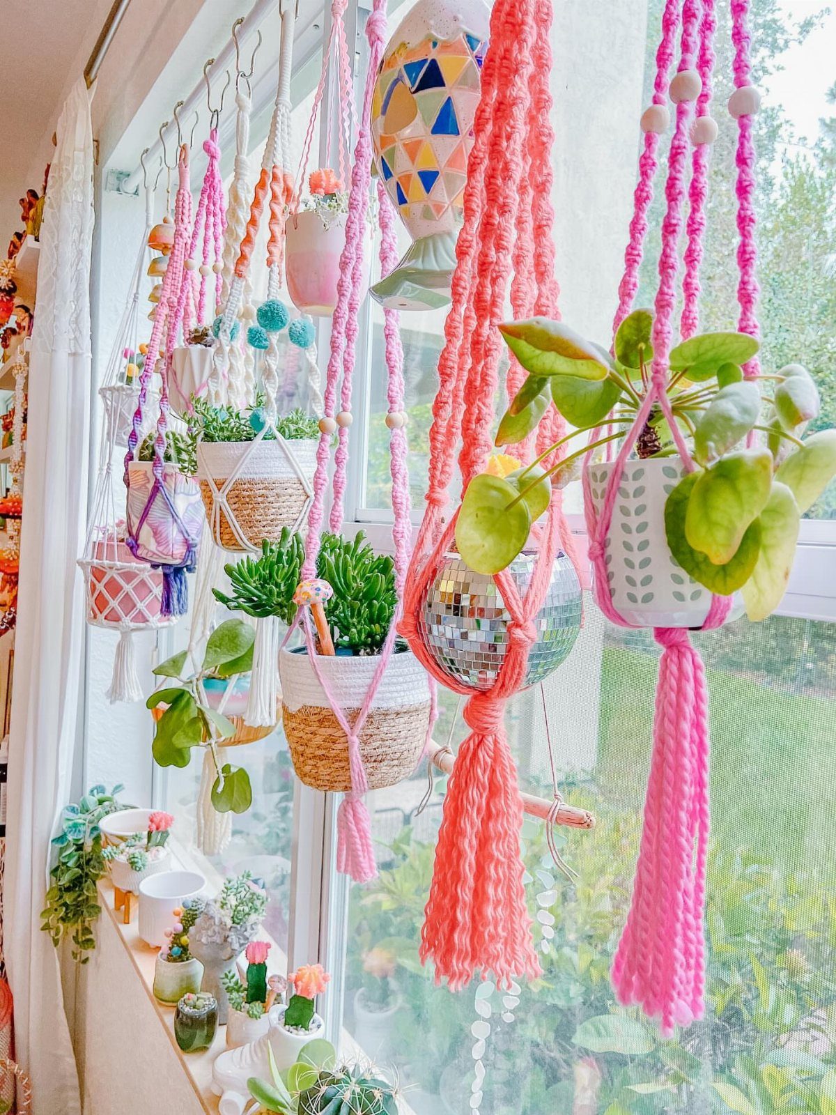 Colorful macramé plant hangers holding green potted plants hanging by a bright window.