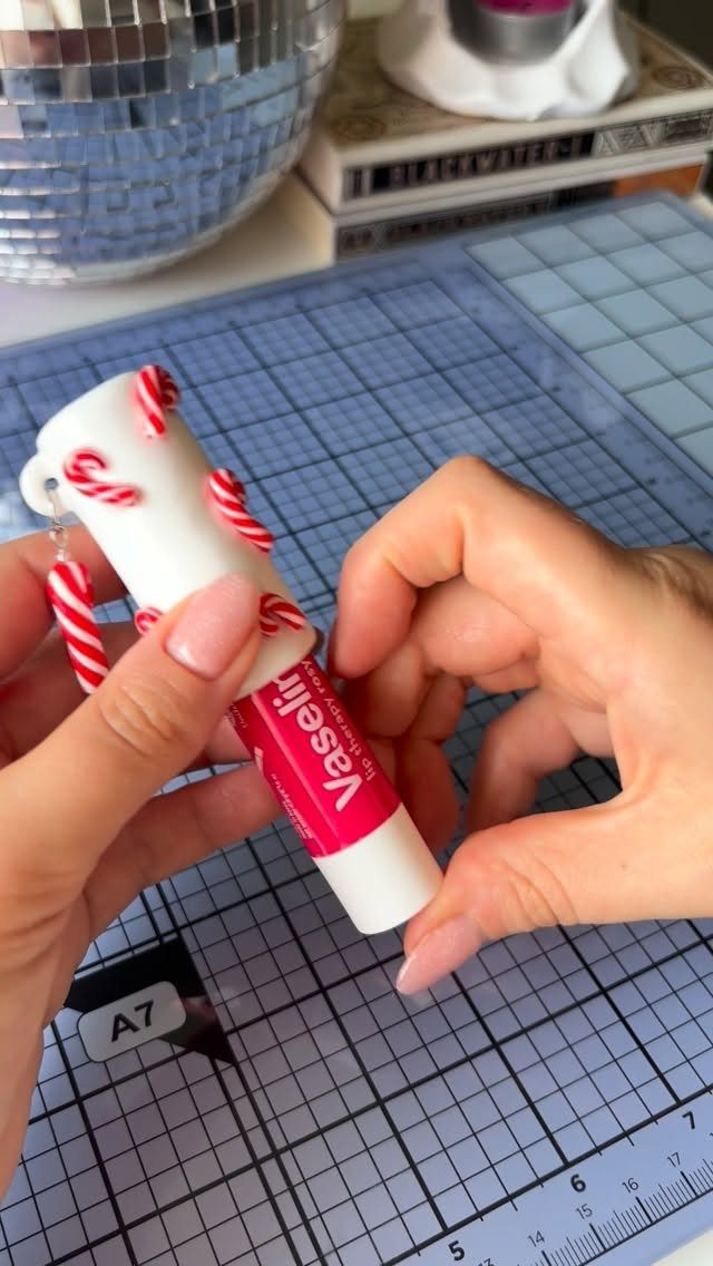 Hands decorating a pink lip balm tube with small candy cane charms on a mat.