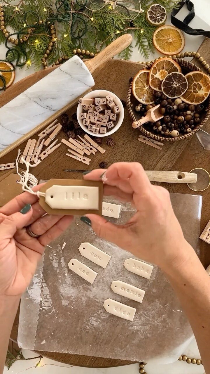Hands holding a stamped white clay gift tag among craft supplies and greenery.