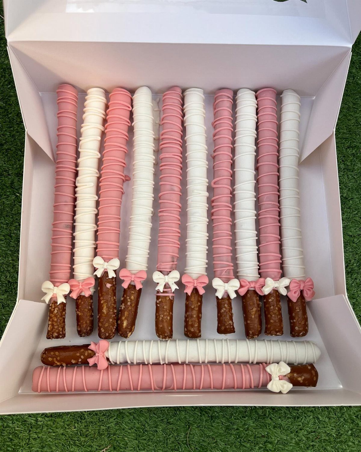 Chocolate-dipped pretzel rods in pink and white with bows in a white box.
