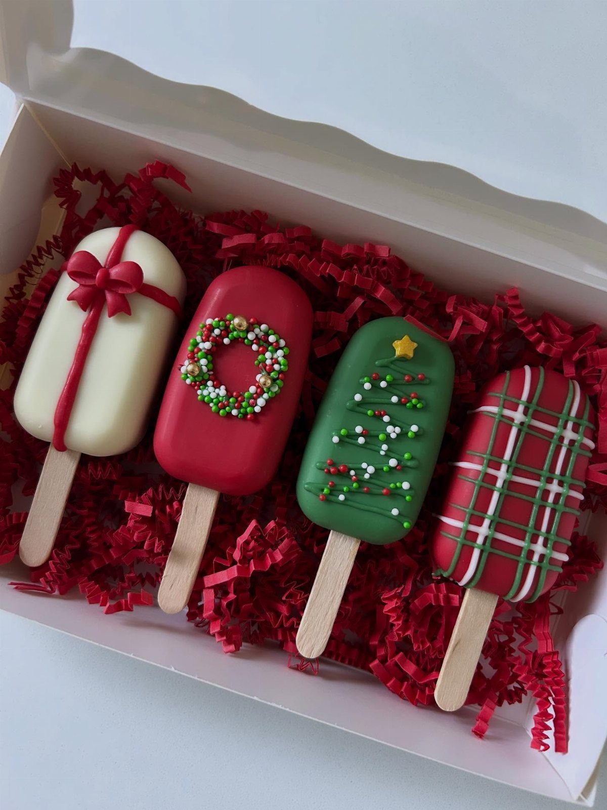 Decorated chocolate cookie pops in a white box with red shredded paper.