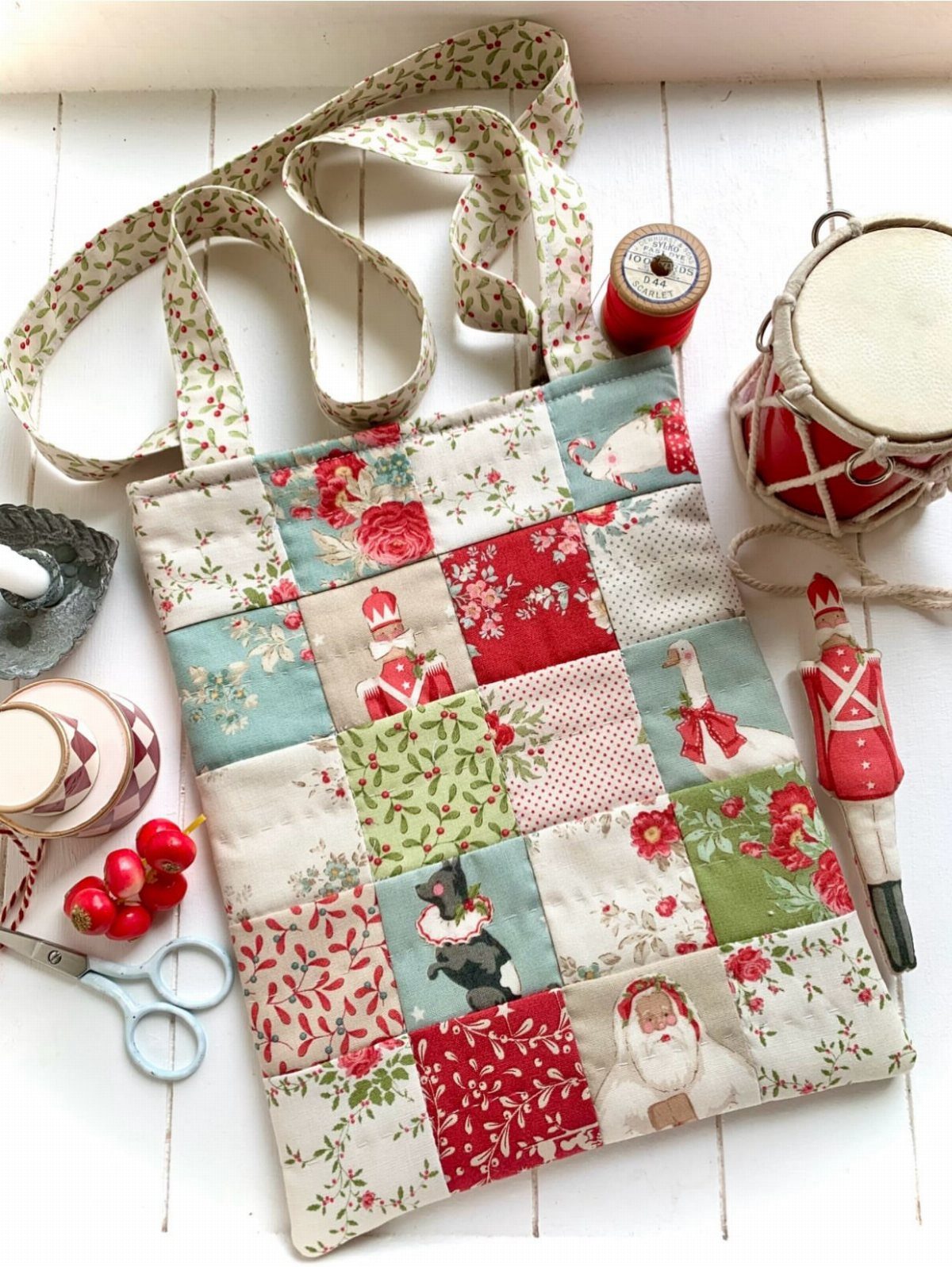 Patchwork fabric tote bag with Christmas patterns surrounded by sewing supplies on white wood.