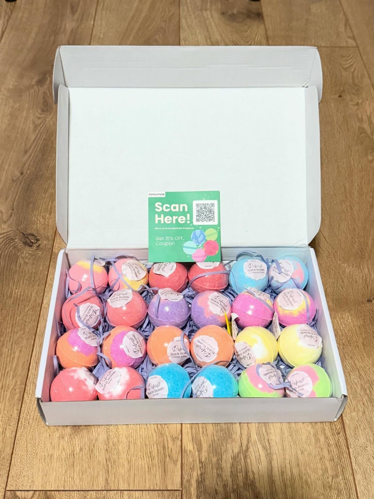 Colorful spherical bath bombs arranged in a white cardboard box on wooden flooring.