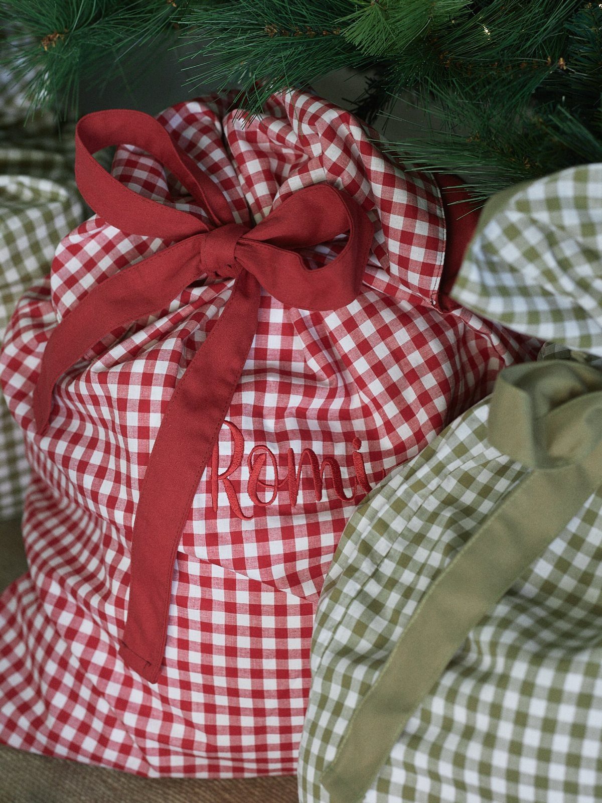 Red and white gingham drawstring gift bags tied with ribbons under pine branches.