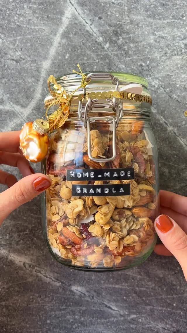Glass jar of homemade granola with a label and gold ribbon held by hands.