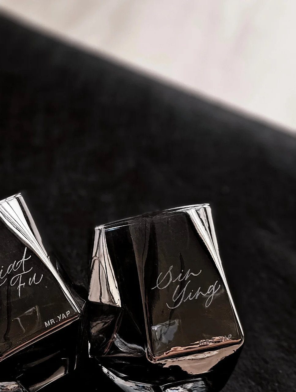 Two black square glasses with etched cursive names on a dark surface.