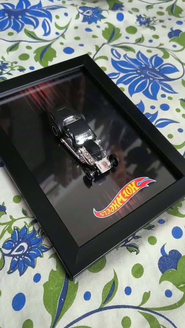 Hot Wheels toy car displayed in a black shadow box frame on floral fabric