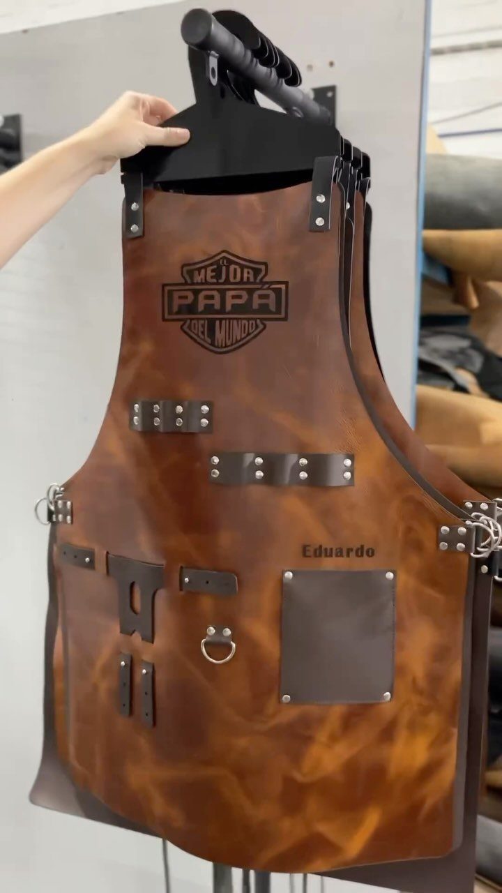 Brown leather personalized apron with pockets and text hanging on a black stand.