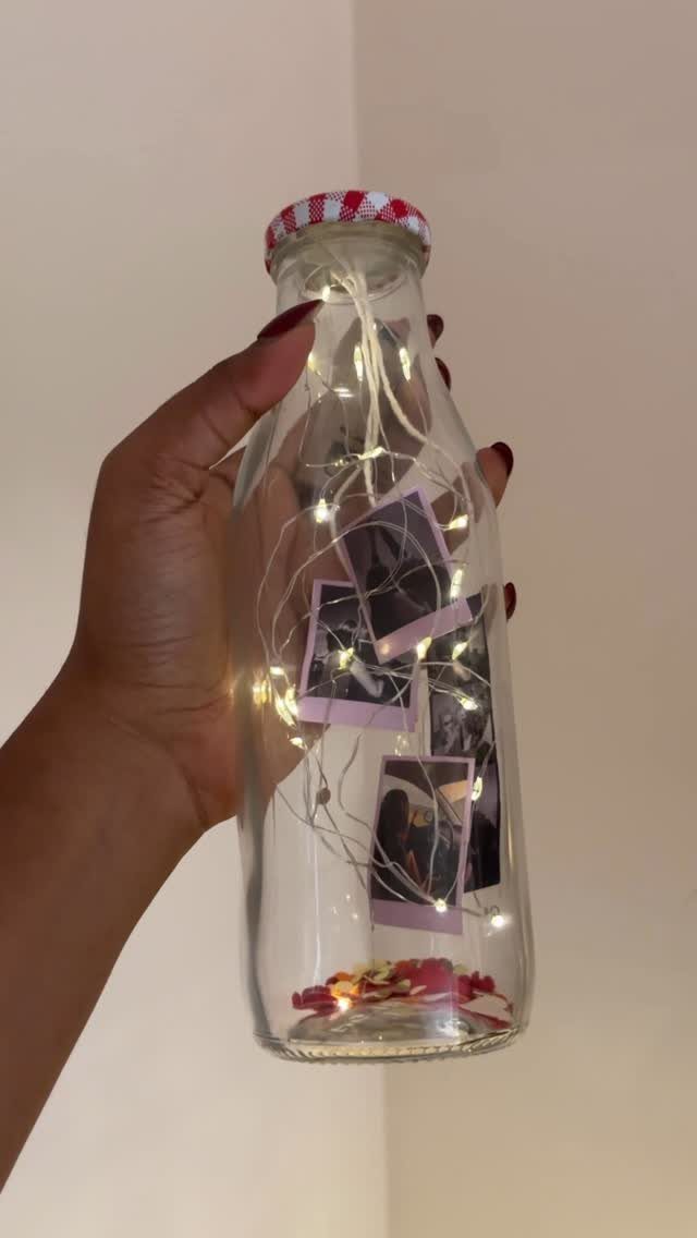 Glass bottle containing polaroid photos, fairy lights, and dried petals held by hand.