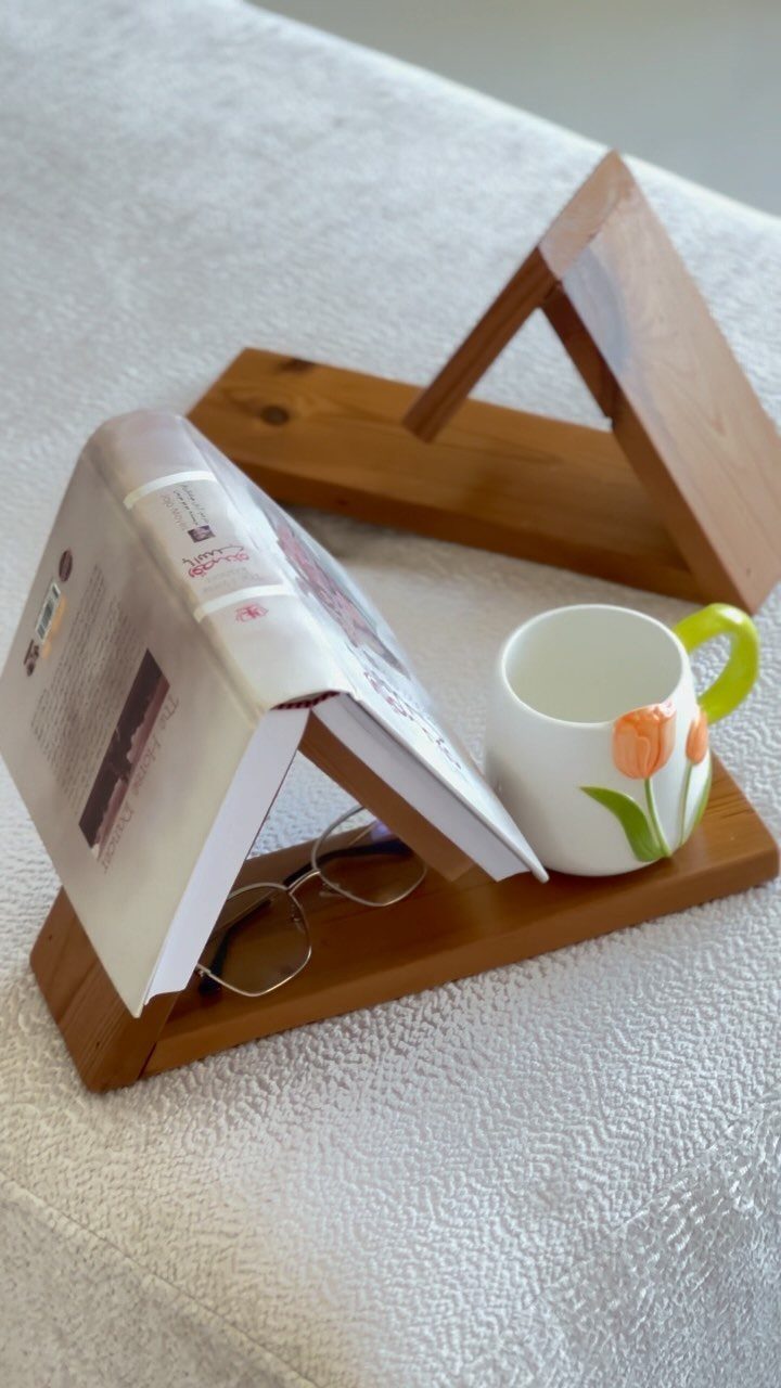 Wooden book stand holding an open book, glasses, and a floral ceramic mug.