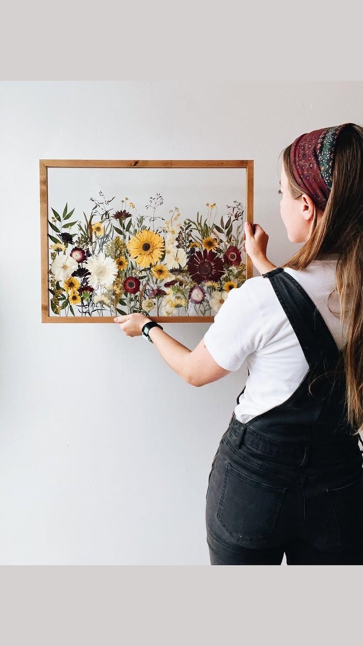 Woman holding a wooden frame with colorful pressed flowers against a white wall