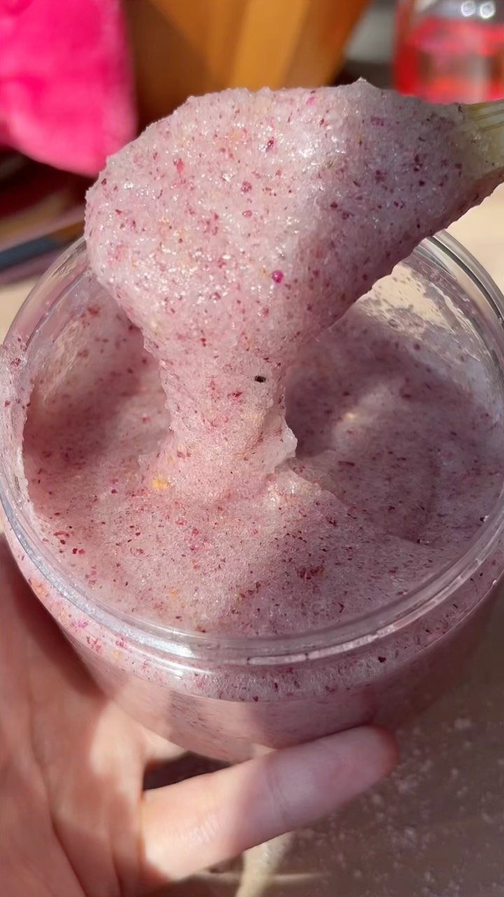 Pinkish-purple sugar scrub being scooped from a clear plastic jar with a spoon