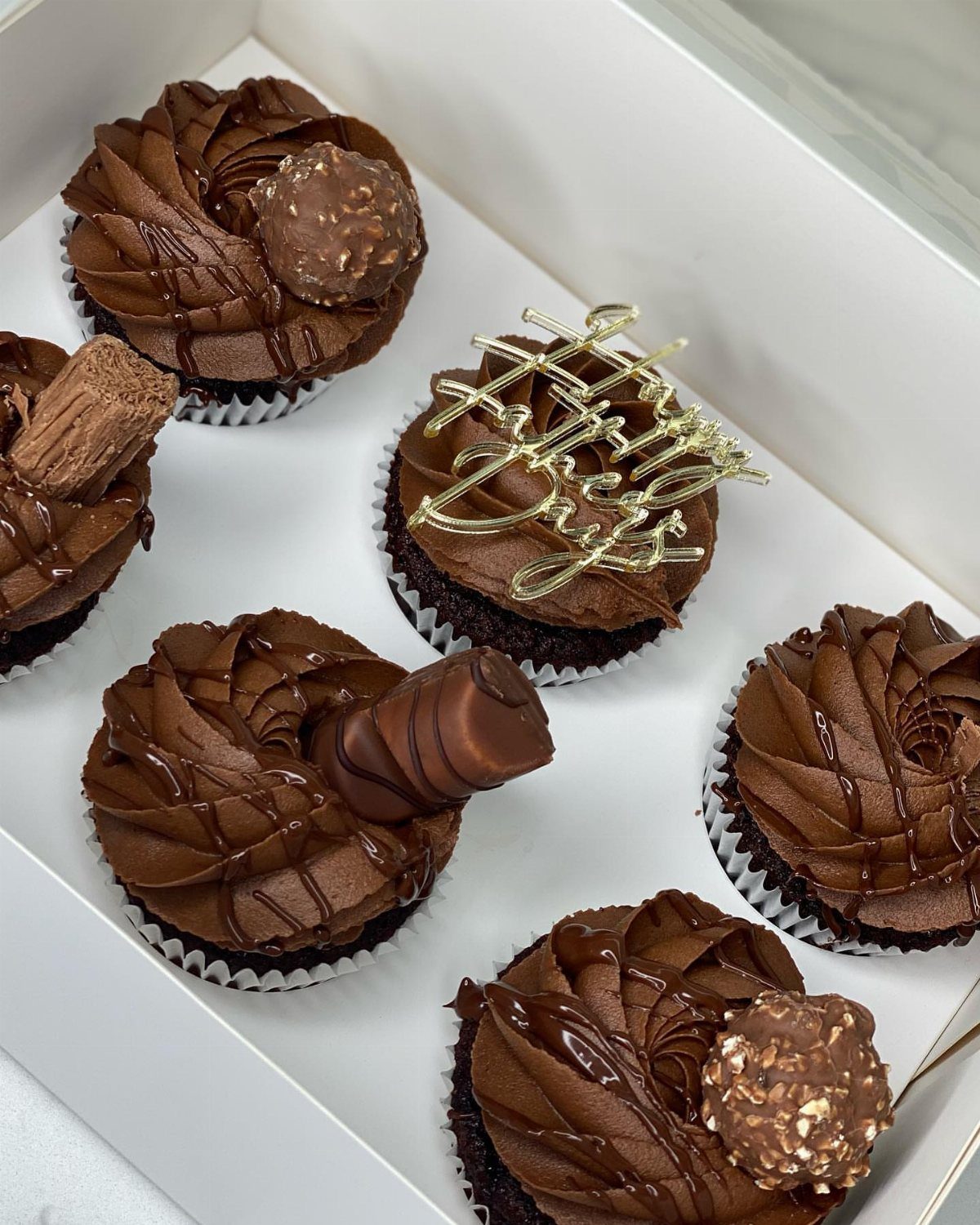 Chocolate cupcakes with frosting and candy toppings in a white gift box.