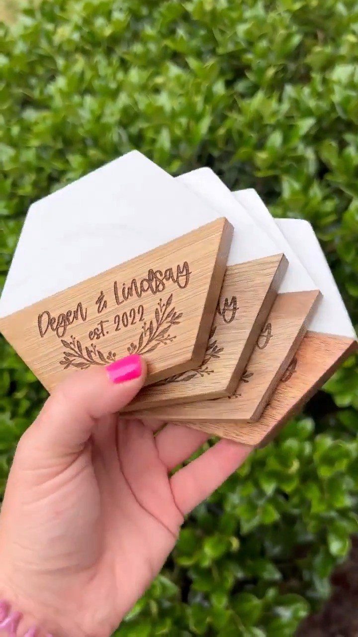 Hexagonal wooden and white coasters engraved with names and a date held outdoors.