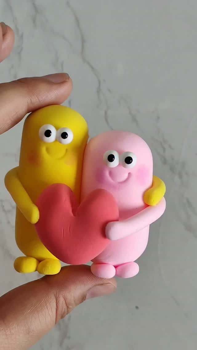 Two small clay figures, yellow and pink, hugging a red heart.