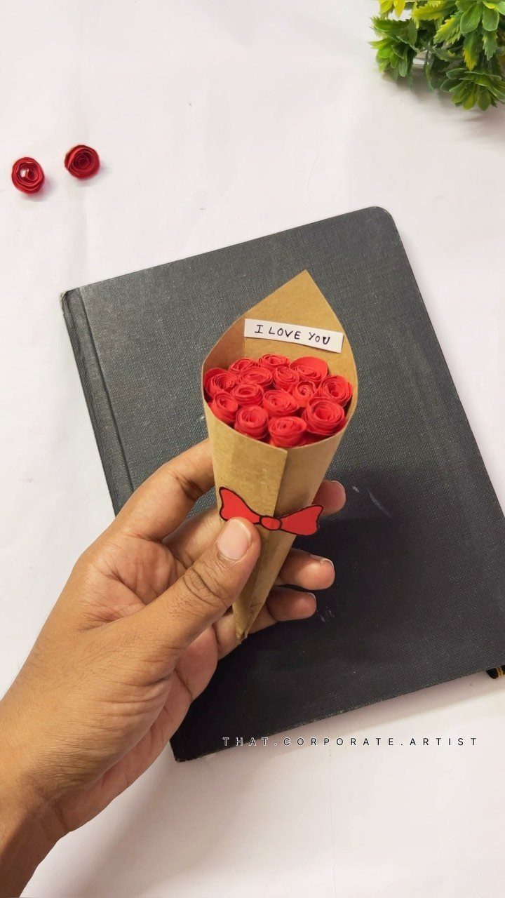 Hand holding a small brown paper cone filled with red paper roses.