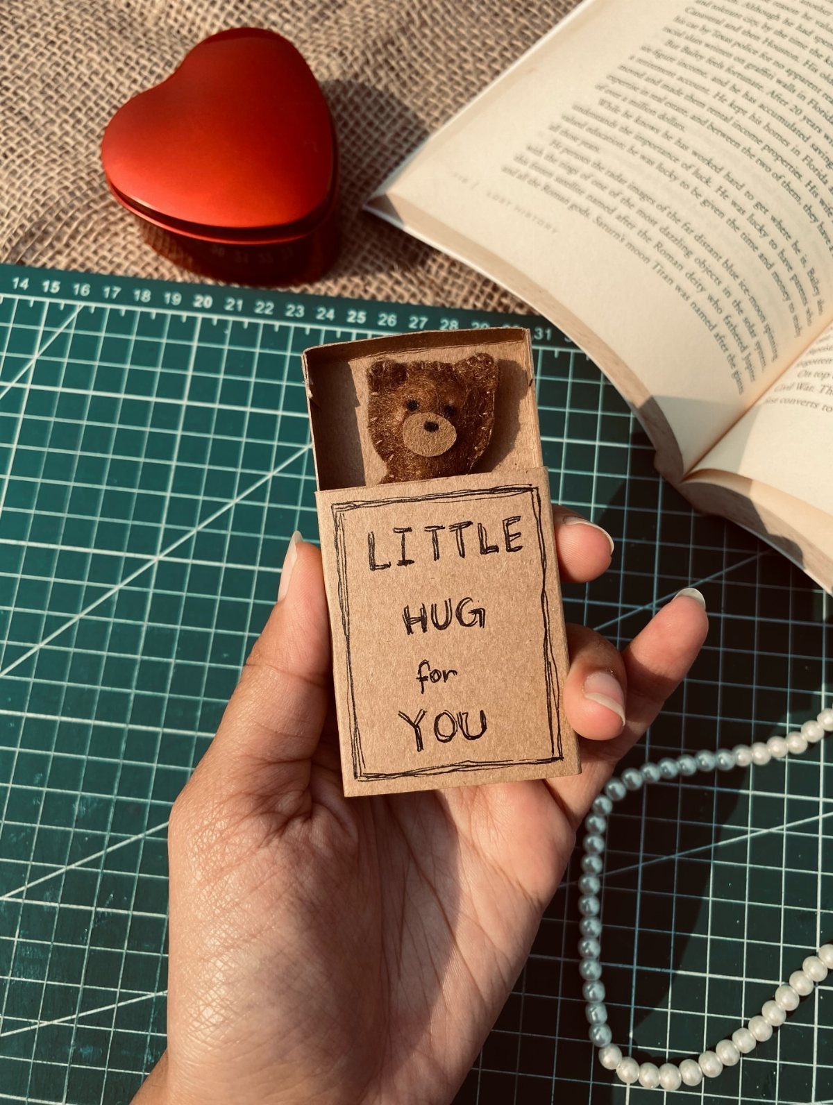 Hand holding a tiny teddy bear in a matchbox labeled Little Hug For You