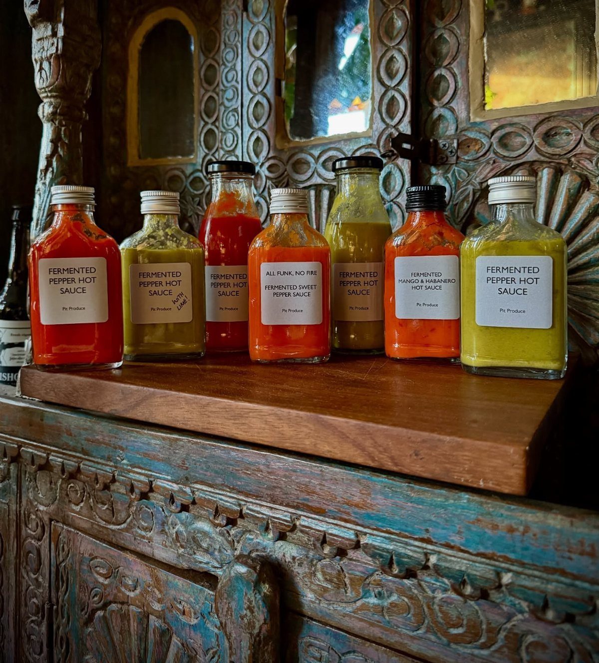 Six bottles of colorful fermented pepper hot sauce on a carved wooden shelf.