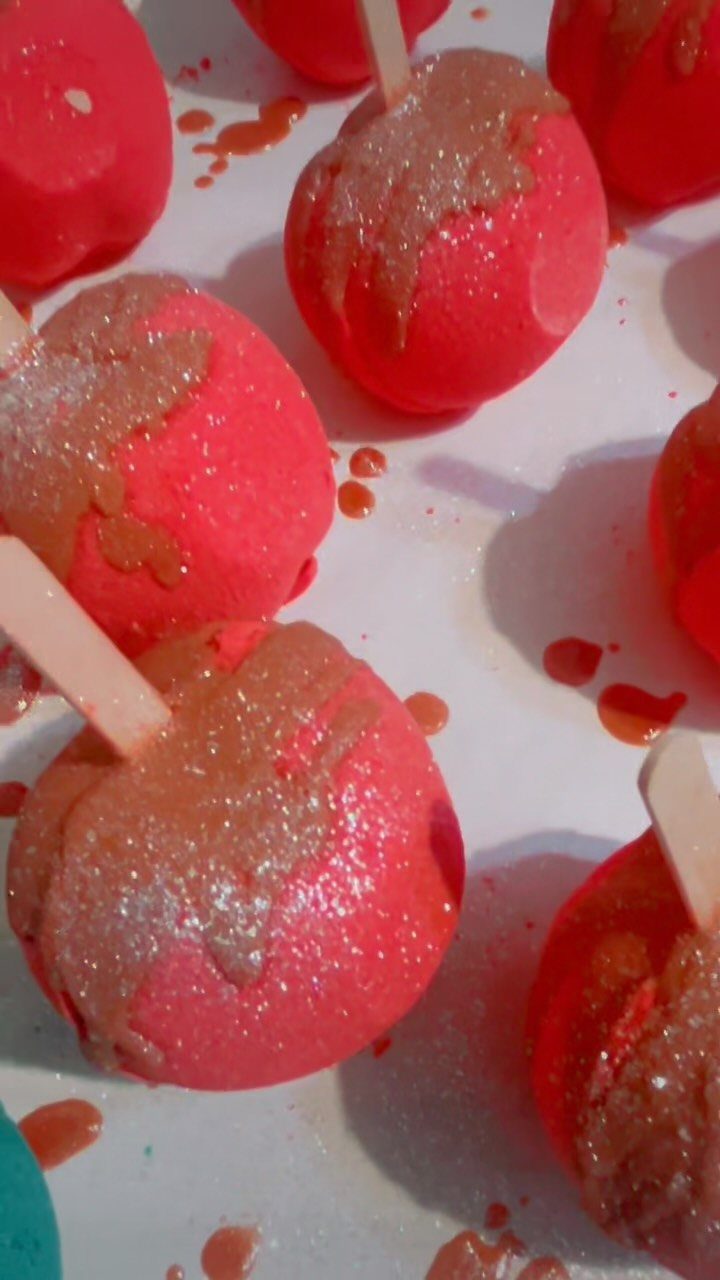 Pink spherical bath bombs with wooden sticks and shimmering glittery topping on white surface
