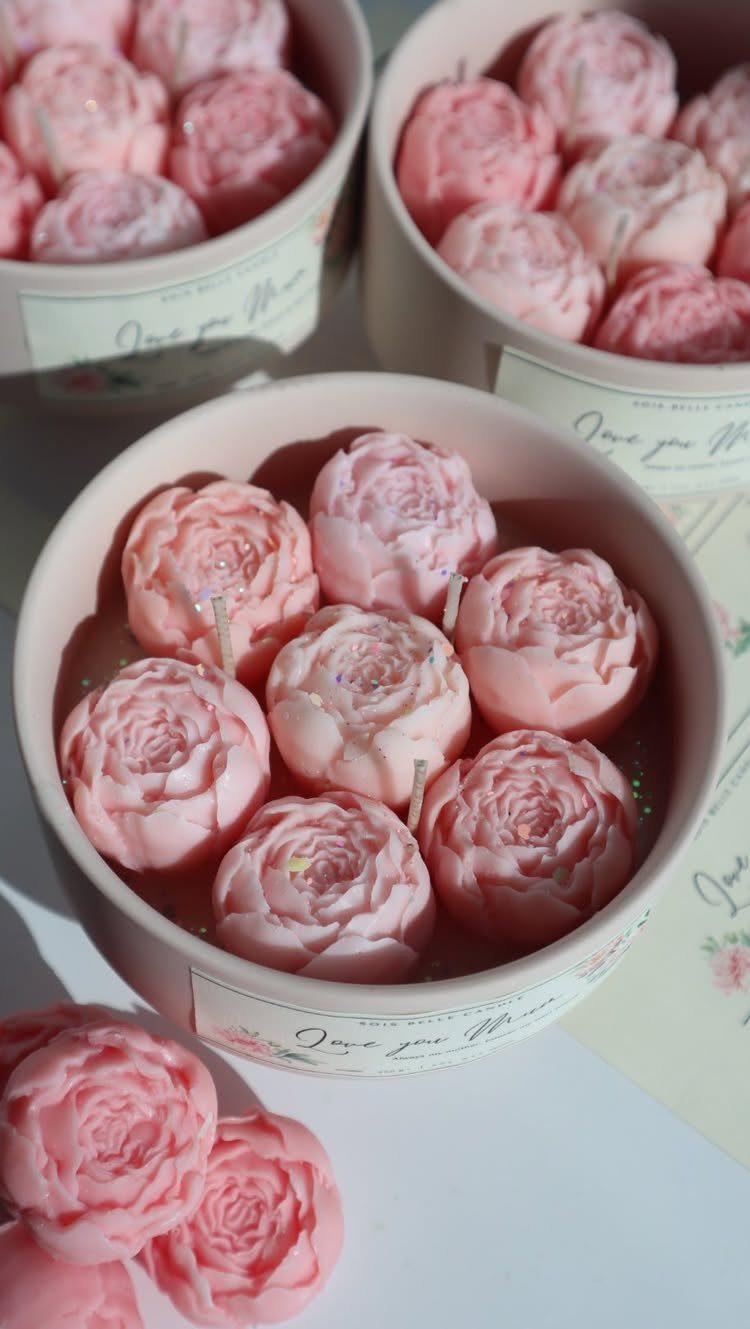 Pink rose-shaped candles in white containers with decorative labels on a white surface.