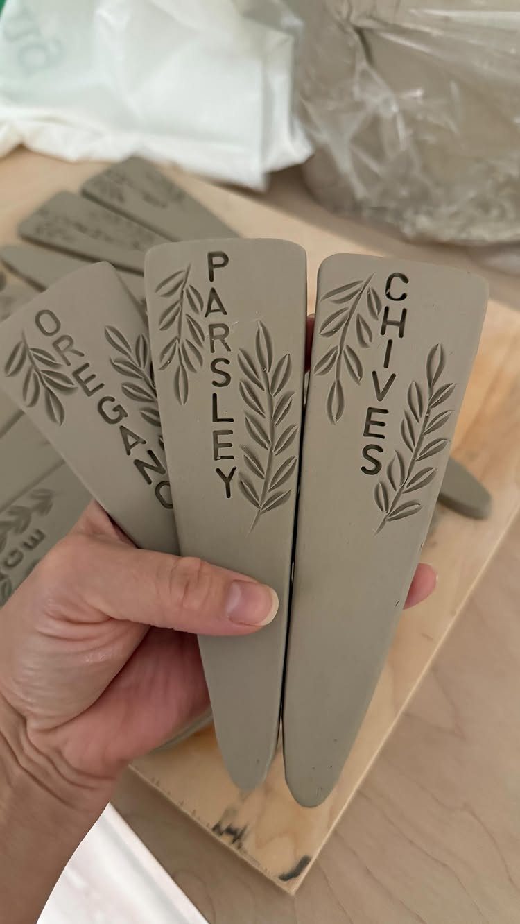 Hand holding three grey clay plant markers stamped with herb names and leaves.