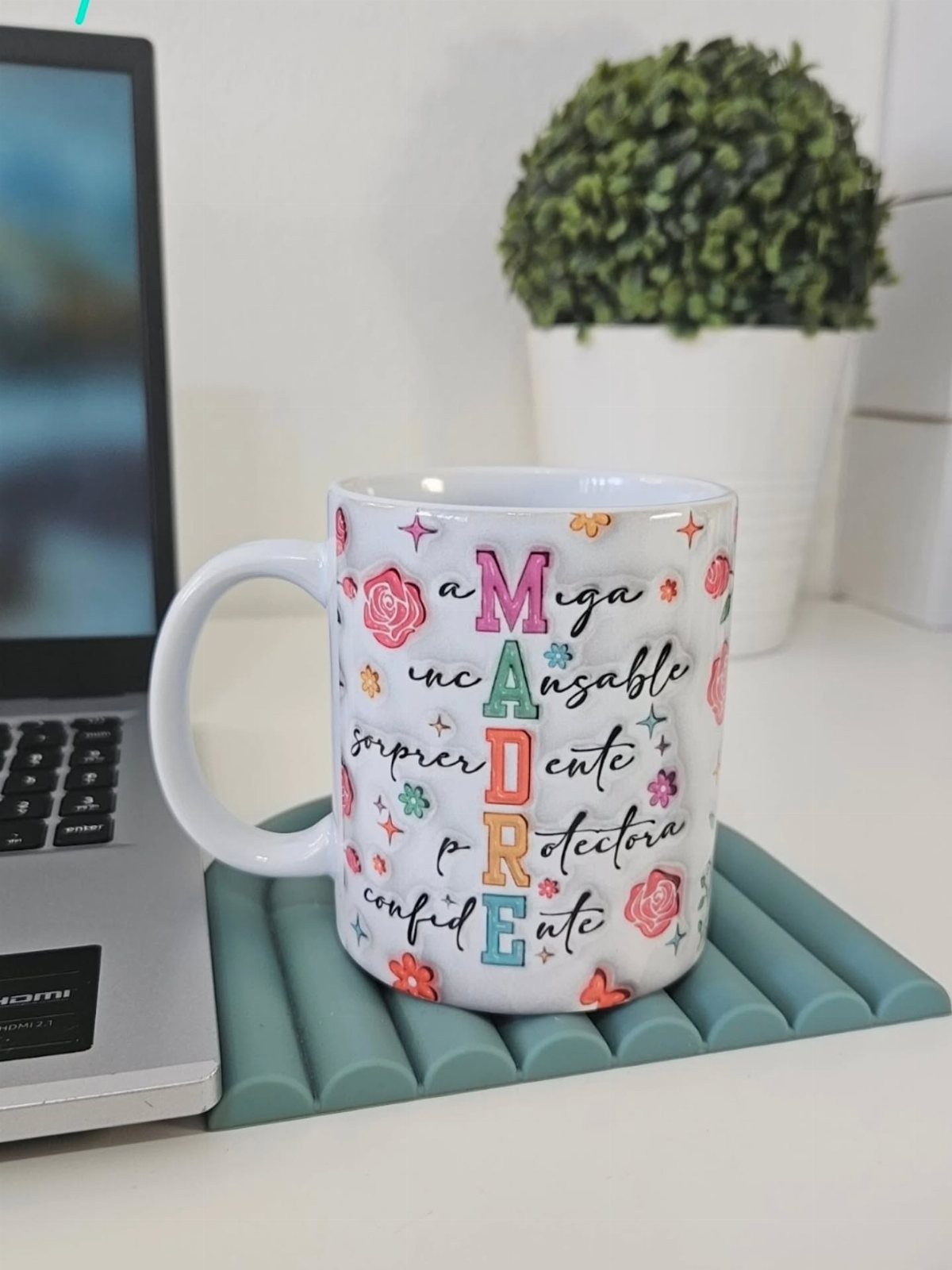 White ceramic mug with colorful floral designs and text on a teal coaster.
