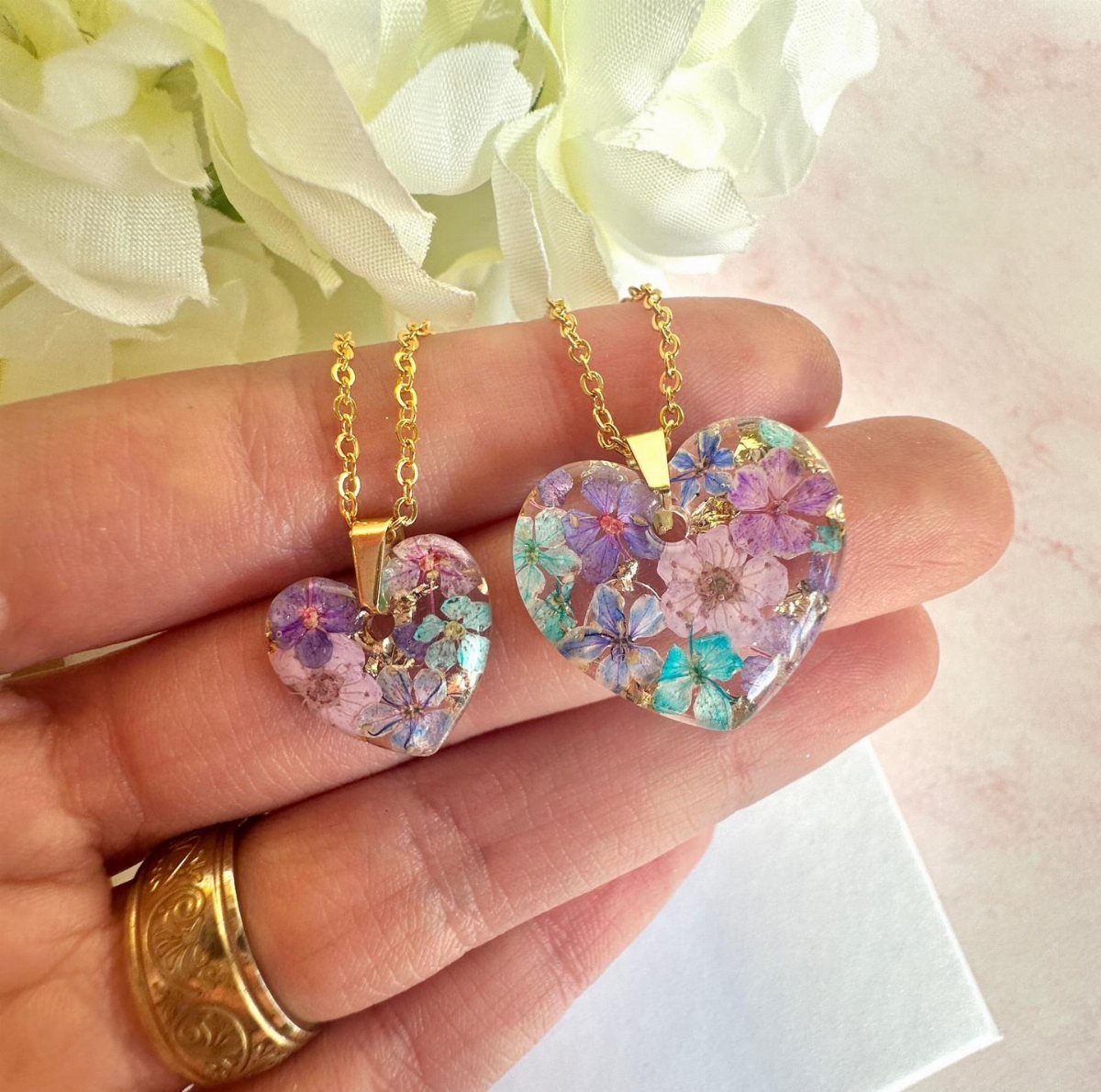 Two heart-shaped resin necklaces with dried flowers held in a person's hand.