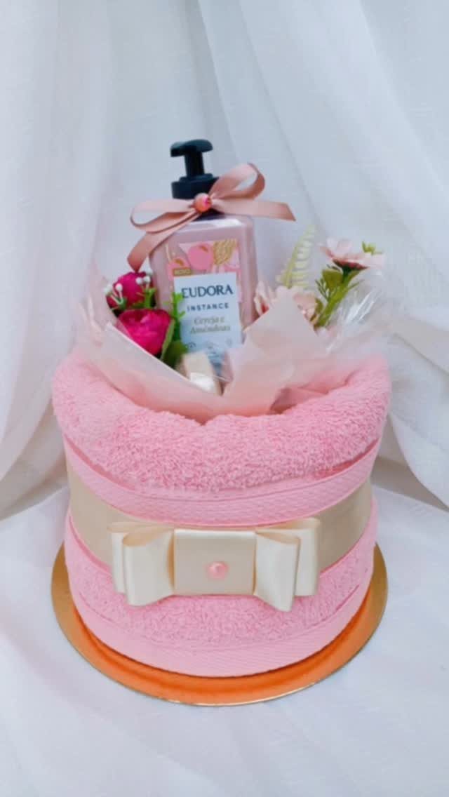 Pink towel cake gift topped with lotion and flowers on a gold base