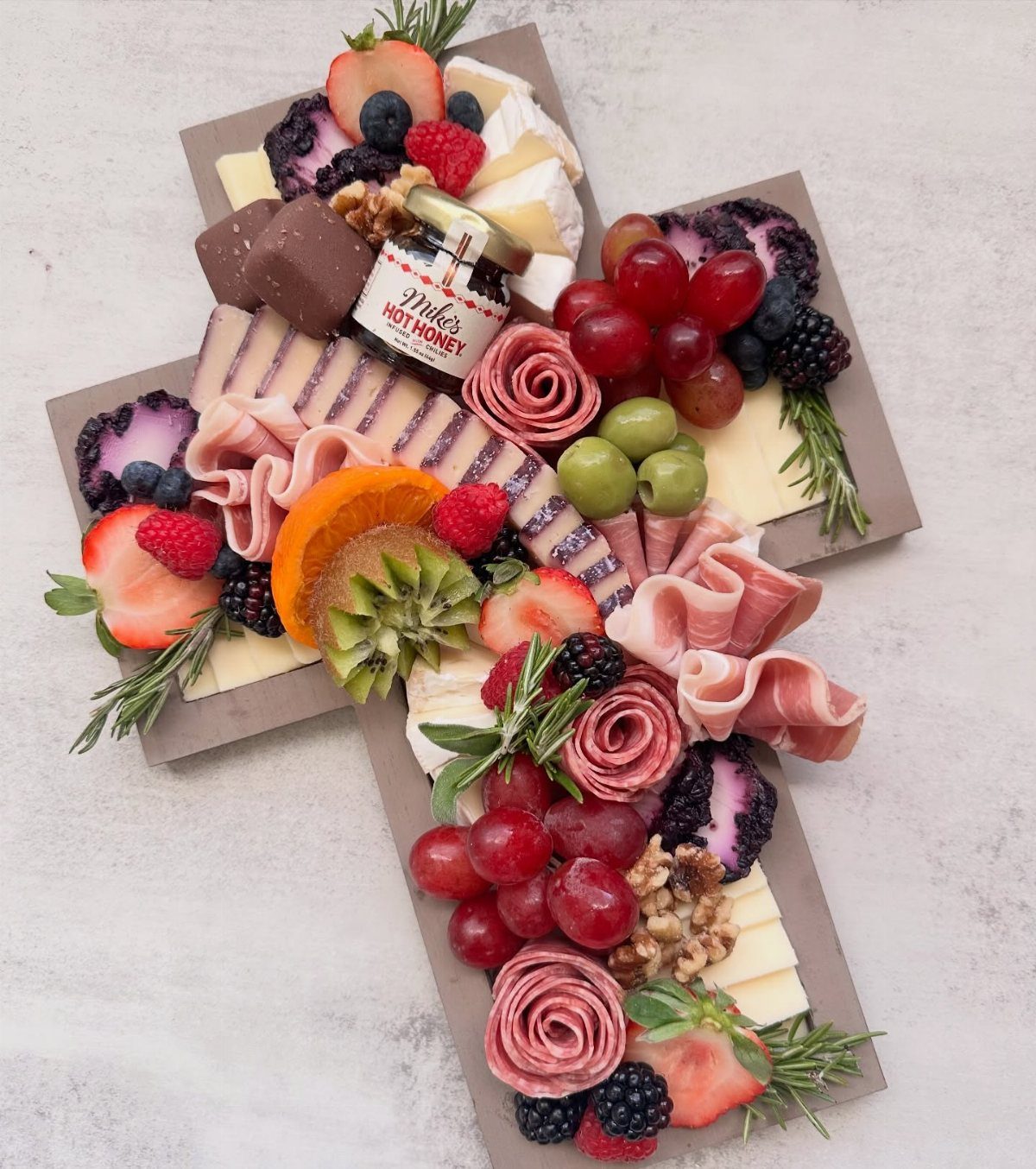 Cross-shaped charcuterie board with meats, cheeses, fresh fruits, and a jar of honey.