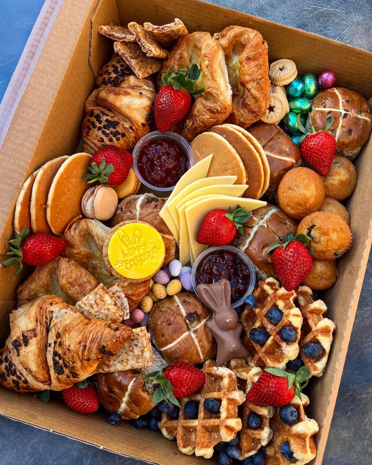 Breakfast board with pancakes, waffles, croissants, and strawberries in a cardboard box.